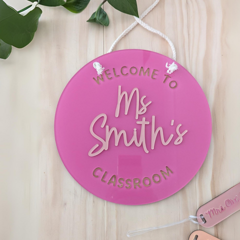 A close-up of a pink, round acrylic classroom sign with the text "Welcome to Ms Smith's Classroom," featuring a white rope for hanging. The sign is a custom gift for teachers, adding a welcoming touch to any classroom