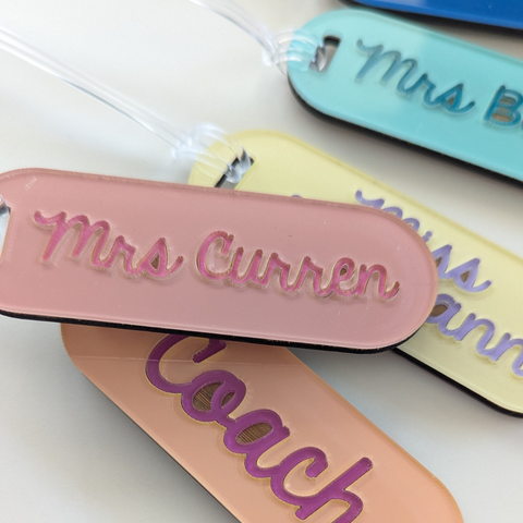 A close-up view of personalized acrylic teacher keyrings with engraved names such as Mrs. Curren and Miss Seigmann. The tags are in peach, teal, and yellow colors with a clear bag tag loop attached.