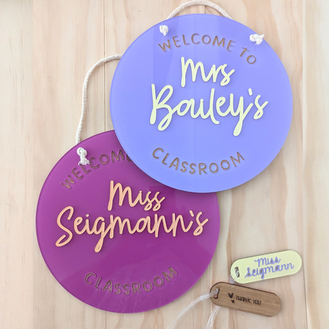 Two personalized acrylic classroom signs in purple and maroon, featuring teacher names "Mrs. Bailey's" and "Miss Seigmann's." The signs have gold-colored text and include a rope for easy hanging, perfect for classroom decor or gifts.