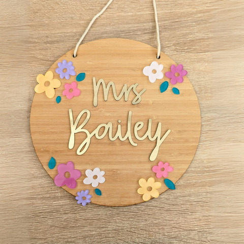 A round bamboo floral name plaque with the name "Mrs. Bailey" in gold acrylic letters. The plaque is decorated with colorful flowers and includes a white hanging cord, displayed on a wooden background.