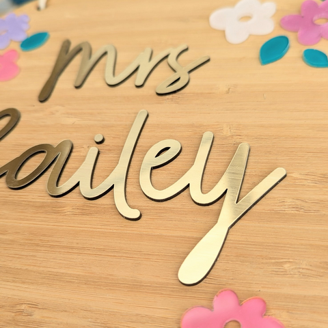 Close-up view of a floral name plaque made from bamboo, featuring the name "Mrs. Bailey" in gold acrylic letters with colorful acrylic flower decorations.