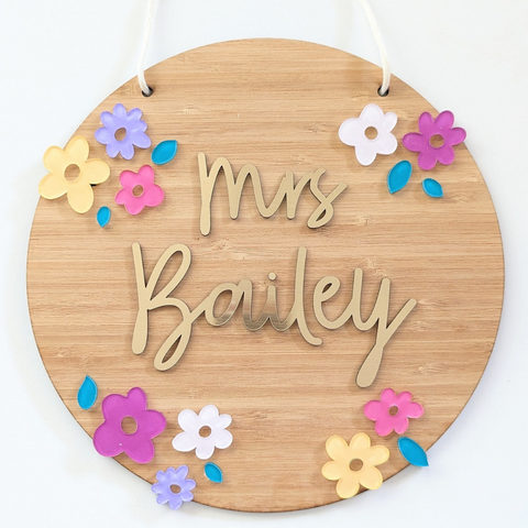 Personalized bamboo name plaque for teachers with colorful acrylic flowers and the name "Mrs. Bailey" in gold acrylic letters. The plaque is round with a soft cord for hanging, perfect for classroom decor.