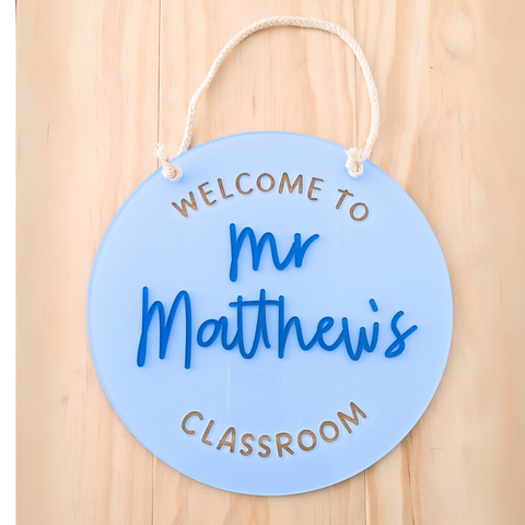 A light blue round acrylic classroom sign with the text "Welcome to Mr Matthew's Classroom," featuring bold blue raised lettering and a white rope for hanging. The sign is placed on a wooden background, perfect for classroom decor or as a gift for teachers.