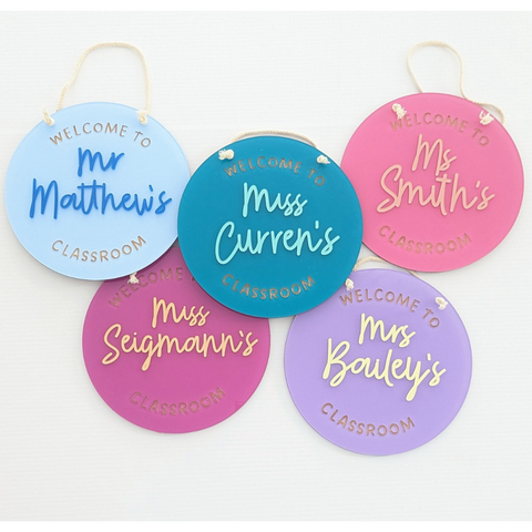 A collection of personalized classroom welcome signs in various colors, featuring teacher names like Mr. Matthew's, Miss Curren's, Ms. Smith's, and Mrs. Bailey's. These round acrylic signs are customized with the teacher's name and "Welcome to [Teacher's Name] Classroom