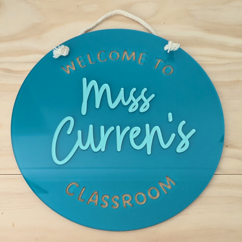 A teal round acrylic classroom sign with the text "Welcome to Miss Curren's Classroom," hanging from a white rope on a wooden background